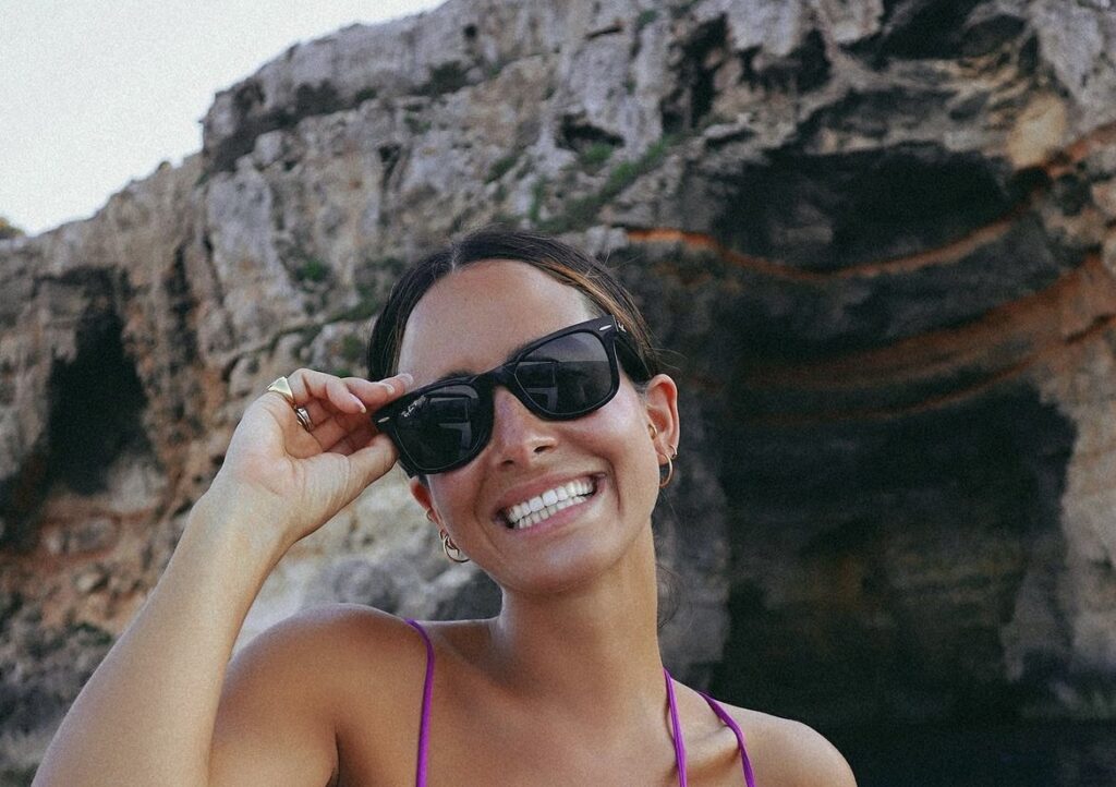 Una mujer tomandose una foto con su traje de baño y sus gafas de sol frente a un muro rocoso