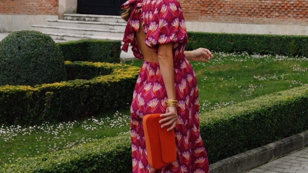 Una mujer caminando en un jardin con un elegante vestido fucsia con topos blancos mientras sostiene su cartera naranja