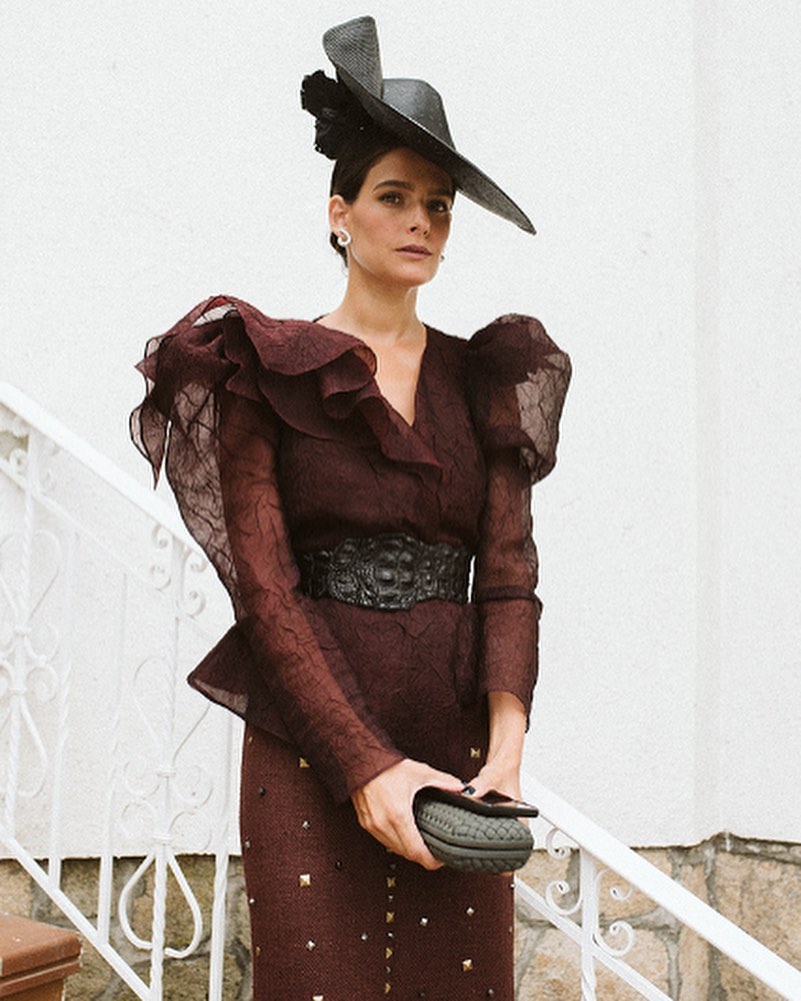 Una mujer baja por las escaleras con un elegante vestido combinado con su sombrero mientras sostiene una cartera