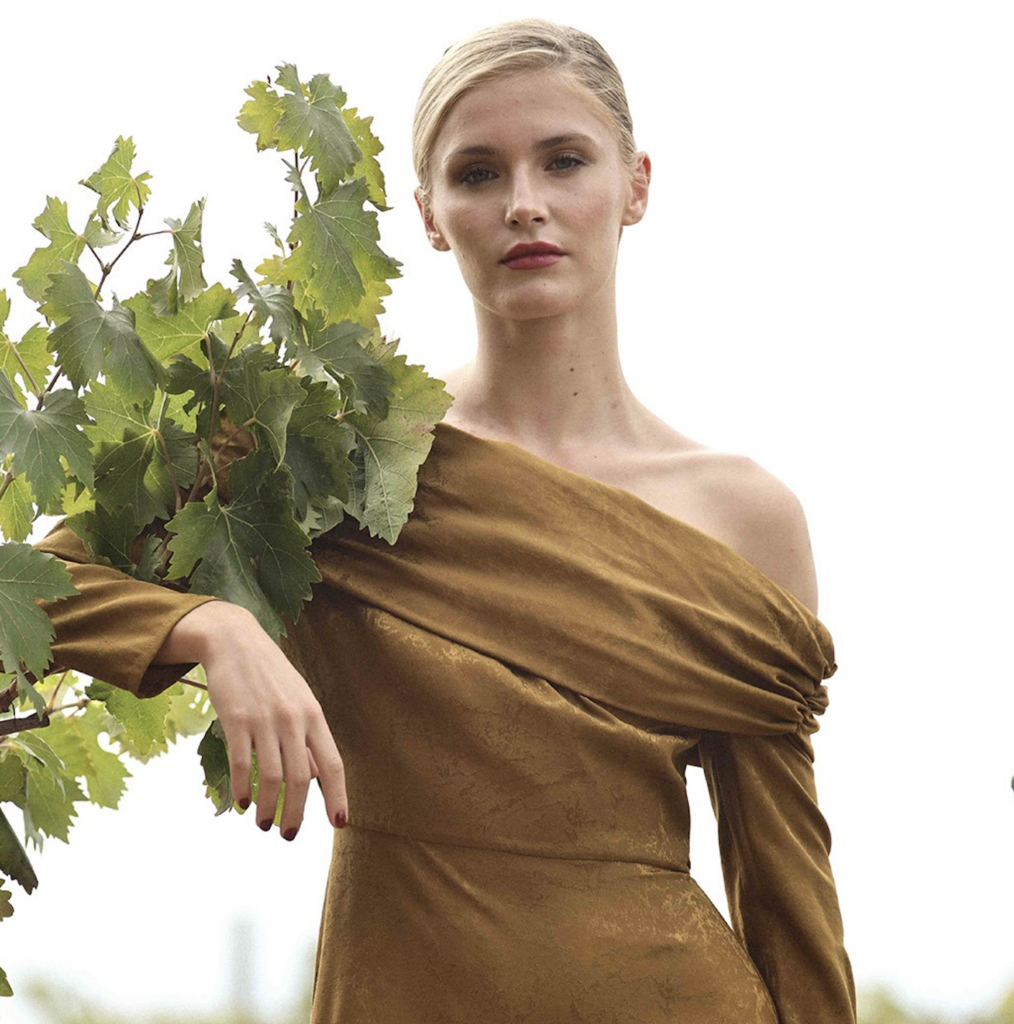 Una mujer con un elegante vestido apoyándose con su brazo derecho una rama de hojas 