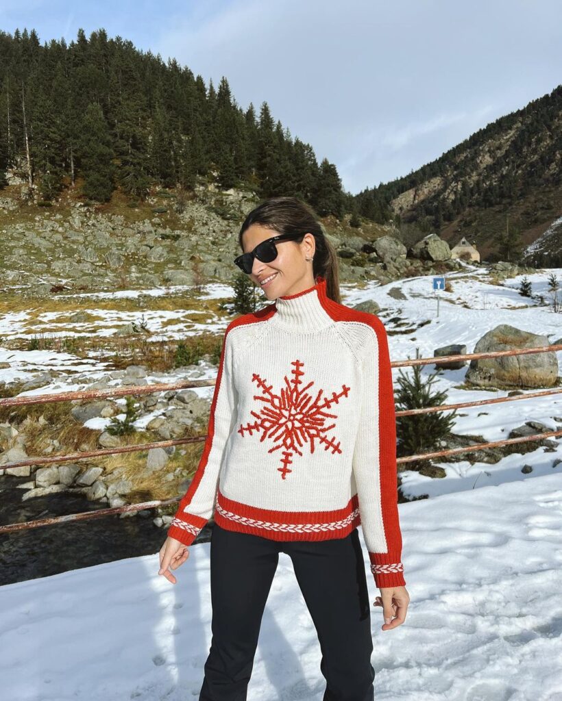 Una mujer usando un abrigo blanco con fraciones rojas en los hombros y un copo de nieve rojo en el pecho